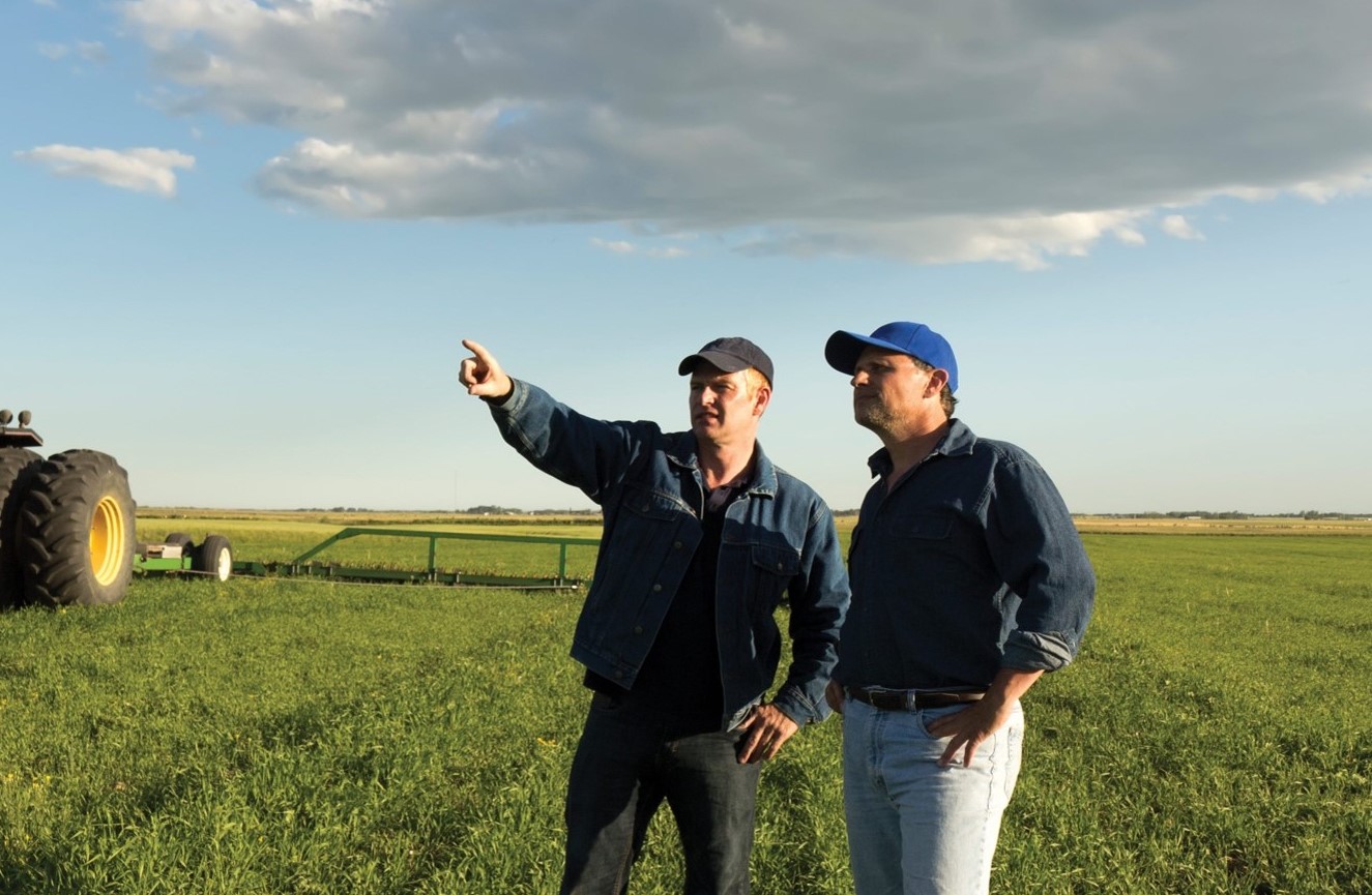 Two people in a farm field with equipment behind them pointing at something in the distance.