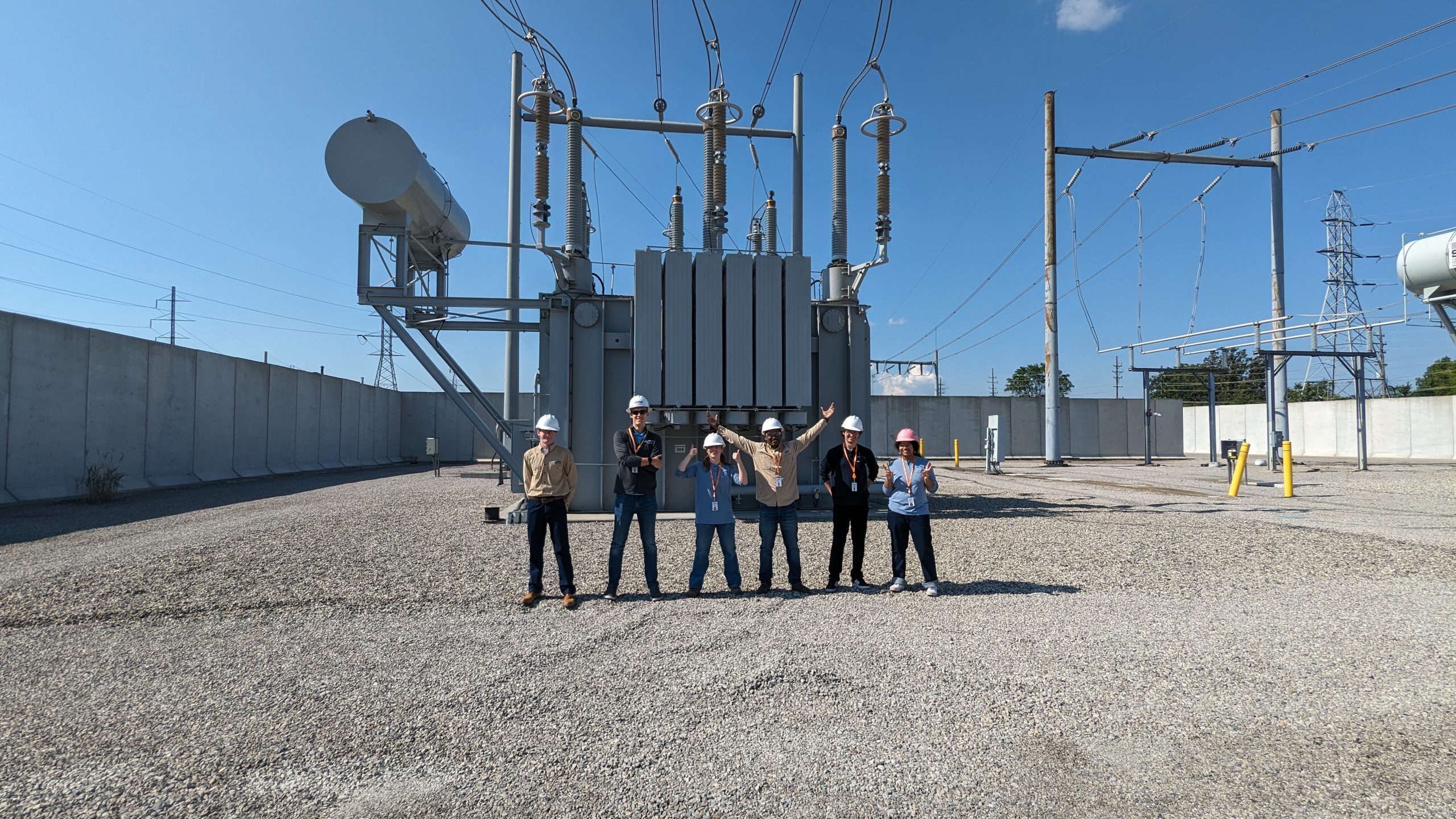 Six young adults in hard hats standing in front of transmission station.