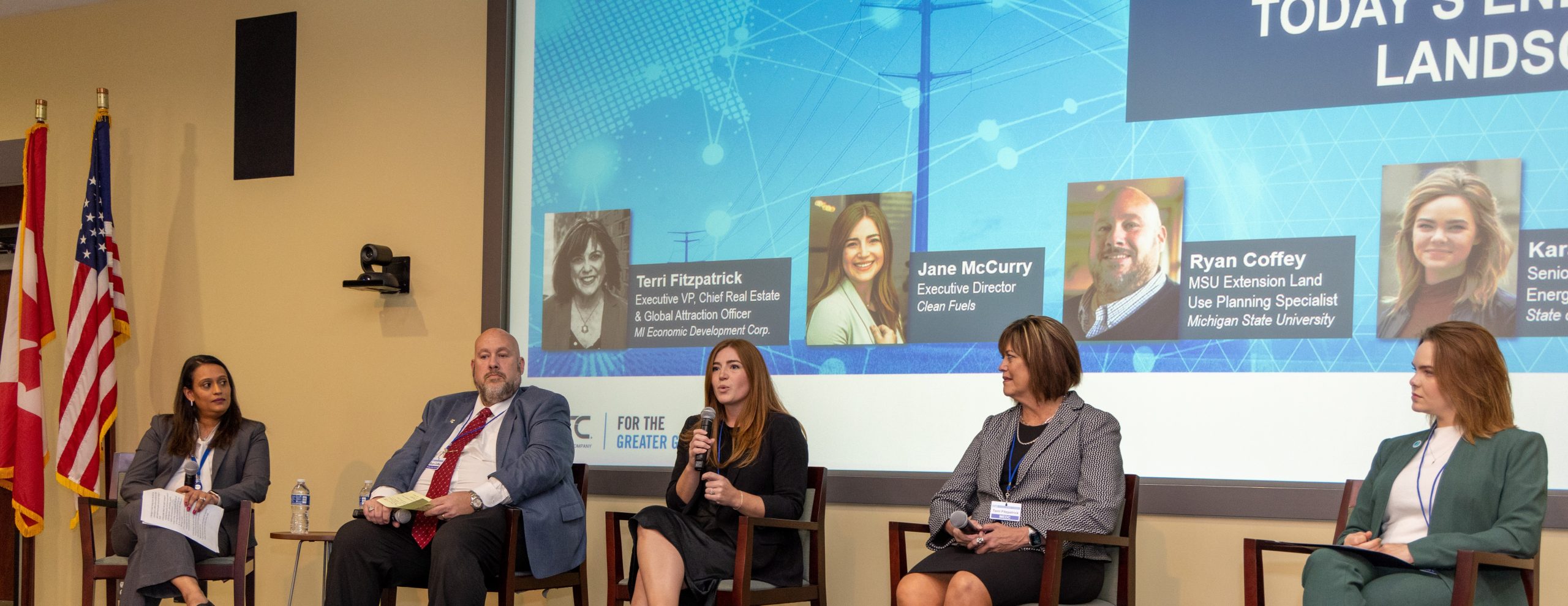 Five people on a stage, third person from the left is holding a microphone and speaking.