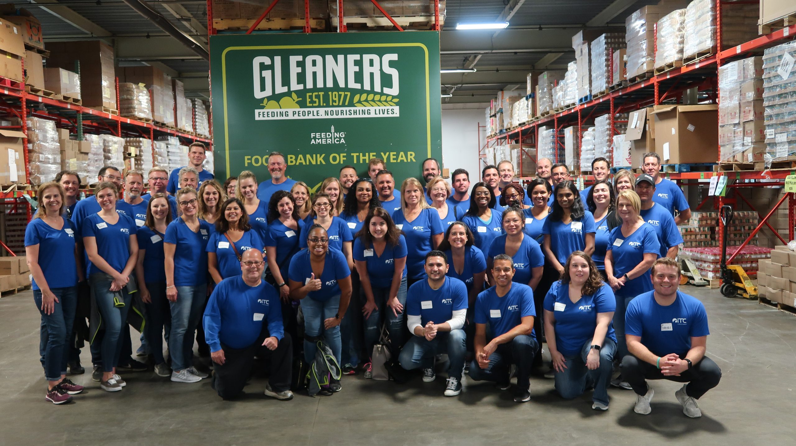Large group of people in blue ITC shirts, in warehouse with Gleaners Food Bank Logo behind them.