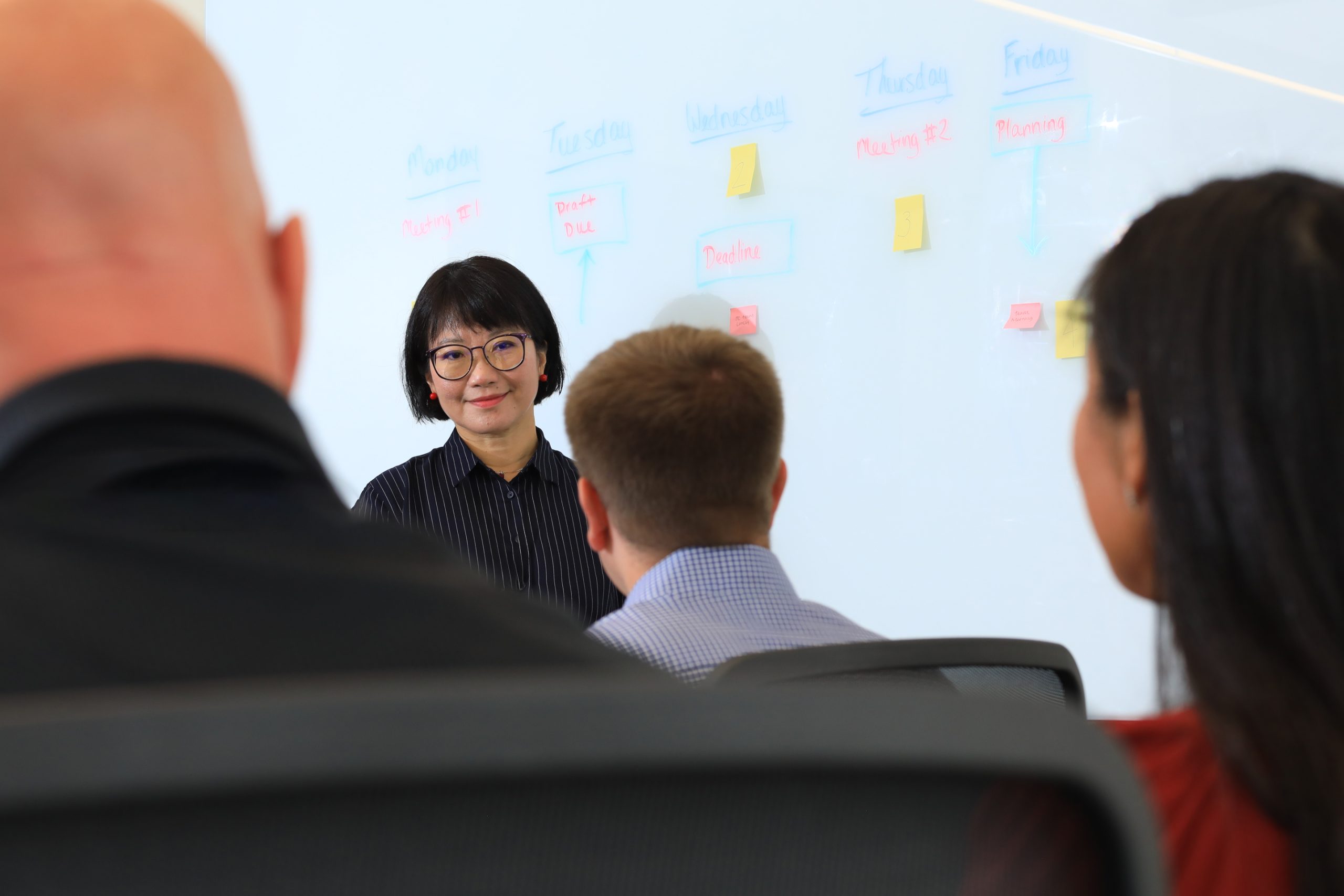 People in meeting room looking at speaker in front of a whiteboard with writing and post-it notes.