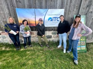 Group of five ITC volunteers standing in front of a fence with an ITC sponsorship banner while holding plants.