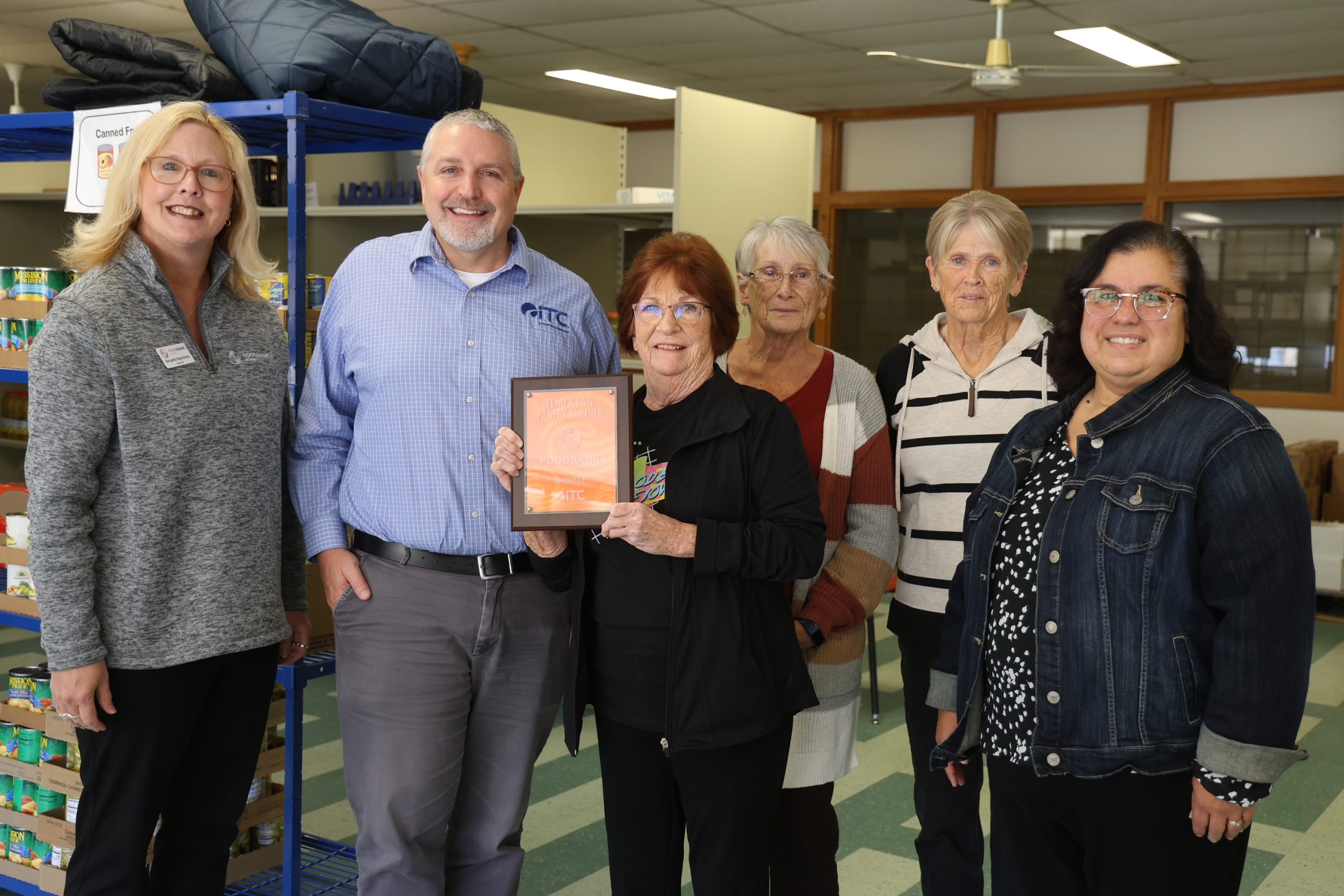 Six people standing in food bank showing a plaque that notes ITCs partnership with the food bank.