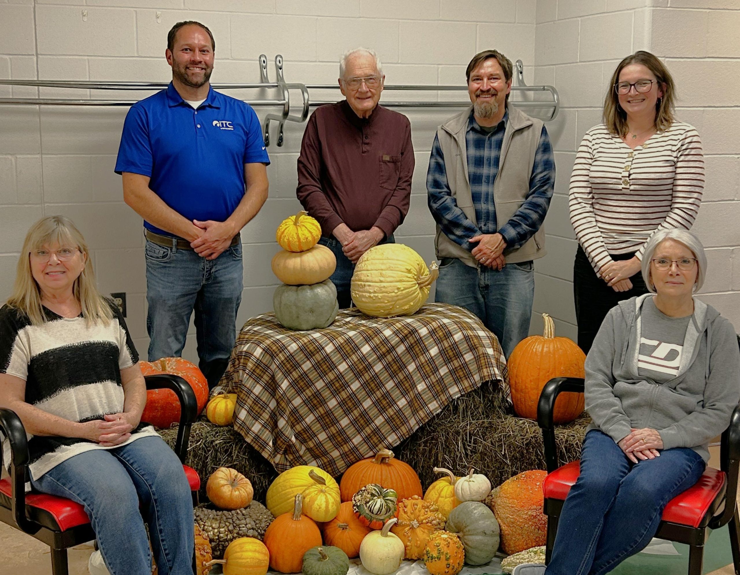 Group of 6 people, some sitting, some standing near a hay bail with pumpkins.