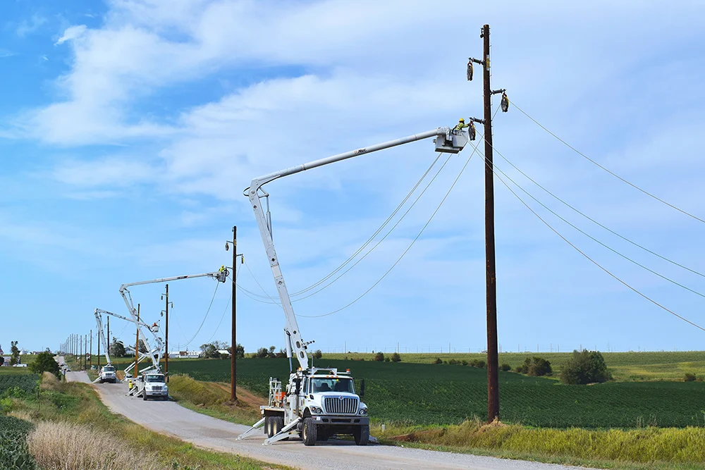 Transmission lines being serviced by workers in trucks alongside a farm road.